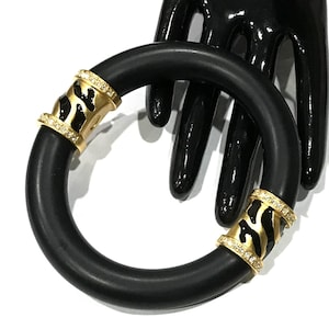 May include: A black bangle bracelet with gold-tone detailing. The bracelet has two gold-tone sections with a black and gold animal print design, set with small, clear crystals. The bracelet is displayed against a black background.