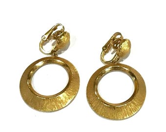 May include: A pair of gold-toned clip-on earrings featuring a circular drop design. Each earring consists of a textured, brushed gold hoop suspended from a matching circular clip mechanism. The metal surface displays a fine, radial brushed finish.