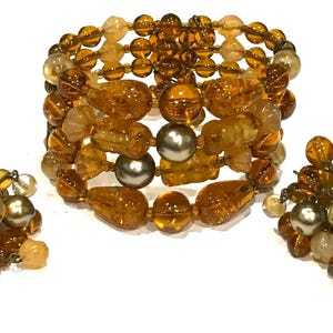 May include: A set of vintage jewelry featuring a wide bracelet and matching earrings. The bracelet and earrings are made of amber-colored beads in various shapes and sizes, with silver-colored accents. The jewelry is displayed on a white surface.