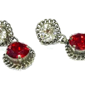 May include: A pair of vintage silver-toned earrings. Each earring features a clear, round gemstone and a dangling, faceted, red gemstone. The gemstones are set in a decorative, twisted metal frame. The earrings are displayed against a white background.
