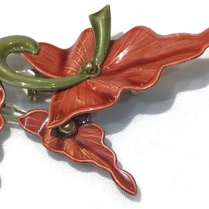 May include: A floral brooch with red leaves and flowers, gold accents, and a green stem. The brooch has a silver-tone pin. The design is reminiscent of a stylized flower arrangement.