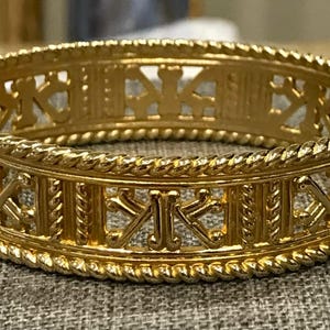 Op de afbeelding: Een goudkleurige scharnierende armband. De armband heeft een open ontwerp met herhaalde geometrische vormen en een touwachtige rand. De sluiting is zichtbaar aan de linkerkant. De armband heeft een diameter van ongeveer 5 cm.