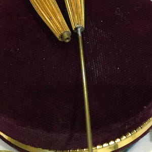 May include: Two gold-toned metal hat pins with elongated, ribbed, spindle-shaped heads. Each pin features a small, white, spherical accent at the very tip. The pins are positioned upright, with one detached from its long, thin, straight metal shaft.