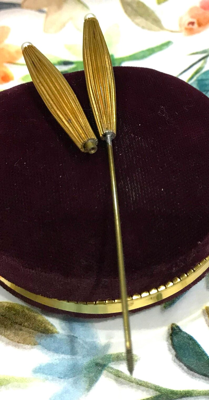 May include: Two gold-toned metal hat pins with elongated, ribbed, spindle-shaped heads. Each pin features a small, white, spherical accent at the very tip. The pins are positioned upright, with one detached from its long, thin, straight metal shaft.