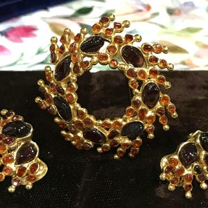 May include: A gold-toned jewelry set consisting of a circular brooch and two matching earrings. Each piece features a textured metal base adorned with numerous small, round, amber-colored stones and larger, smooth, dark brown, teardrop-shaped stones.