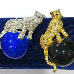 May include: Two leopard brooches, one silver-toned with clear crystals and blue accents perched on a blue sphere, and one gold-toned with clear crystals perched on a black sphere. Both feature detailed feline figures with long tails and sparkling bodies.
