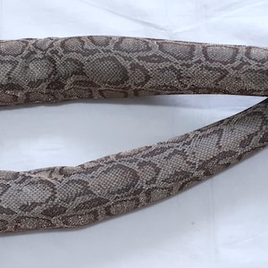 May include: Two arm sleeves featuring a snakeskin design in brown and beige tones. These fingerless sleeves cover the arm from the wrist to the upper arm. The material has a textured finish, resembling snake scales.