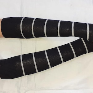 Puede incluir: Calentadores de brazo negros con rayas verticales blancas. Los calentadores de brazo sin dedos cubren la muñeca y el antebrazo. La tela parece ser de punto. Los calentadores de brazo se muestran en un maniquí.