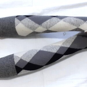 May include: A pair of grey and white argyle pattern arm warmers. The arm warmers feature a thumb hole and are made of a soft material. The argyle pattern consists of black, grey, and white diamonds.