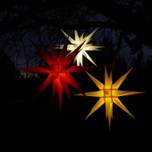 May include: Three illuminated star-shaped lanterns hanging from tree branches. The lanterns are red, white, and yellow.