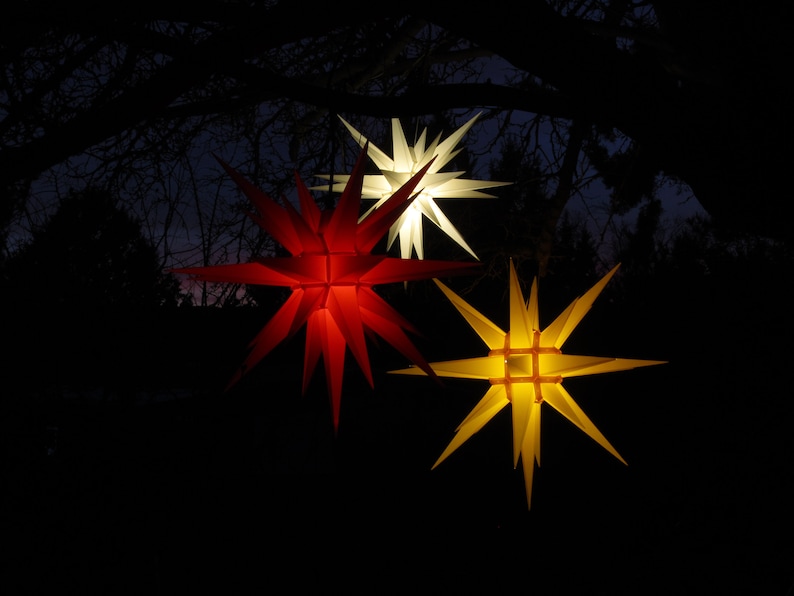 May include: Three illuminated star-shaped lanterns hanging from tree branches. The lanterns are red, white, and yellow.