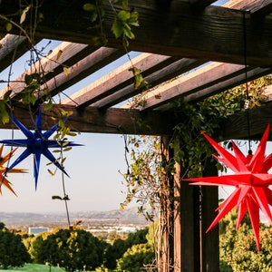 May include: Three paper star lanterns hanging from a wooden pergola. The stars are red, blue, and yellow.