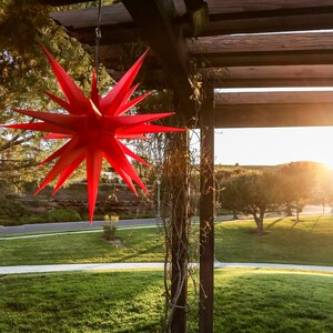 May include: A red star-shaped paper lantern hangs from a wooden pergola with vines growing up the support beams. The lantern is in focus, while the background is blurred and shows a grassy area with trees in the distance.