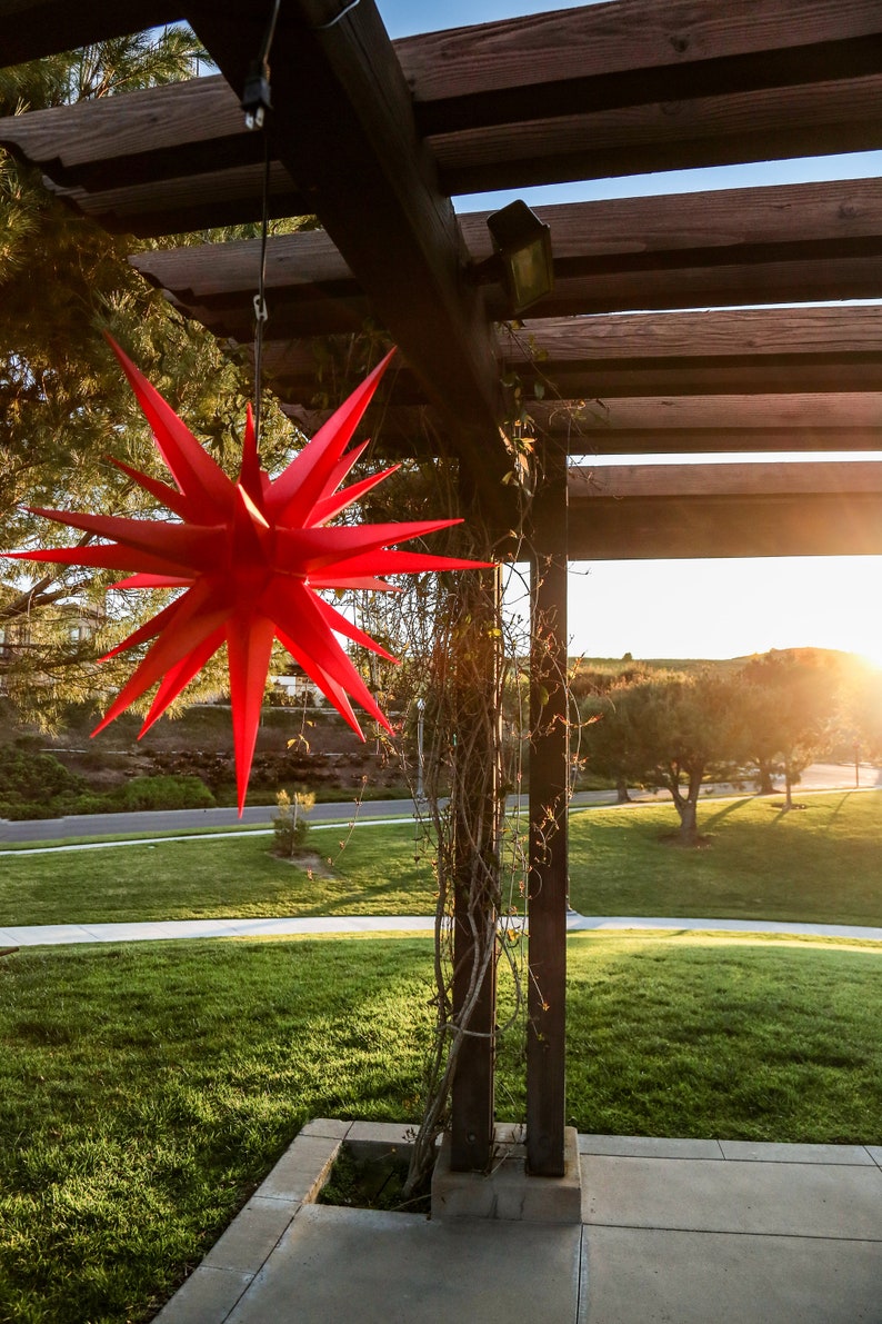 May include: A red star-shaped paper lantern hangs from a wooden pergola with vines growing up the support beams. The lantern is in focus, while the background is blurred and shows a grassy area with trees in the distance.