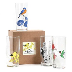 May include: Set of four clear glass tumblers with colorful bird illustrations. Each glass features a different bird species, including a bluebird, a goldfinch, a woodpecker, and a hummingbird. The glasses are packaged in a brown box with the text "One Mercantile" printed on the side.