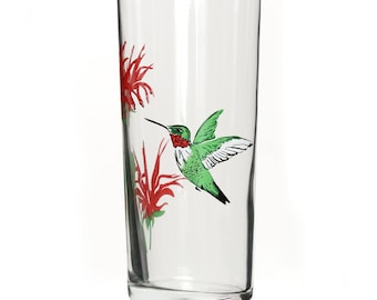 Puede incluir: Un vaso de beber de vidrio transparente con un colibrí y flores rojas pintadas en el lateral.