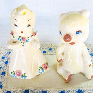 Peut inclure: Un ensemble de figurines en porcelaine vintage représentant une fille en robe blanche avec des fleurs roses et bleues et un ours blanc aux yeux bleus et au nez rose. Les figurines sont posées sur un napperon en dentelle blanche avec une bordure bleue.