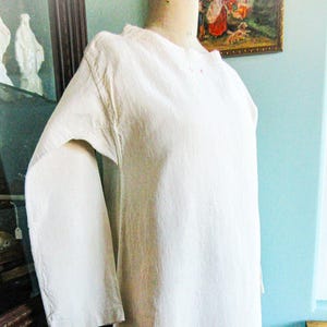 May include: A long-sleeved, off-white dress displayed on a mannequin. The dress has a simple, modest design with a round neckline and a loose fit. The fabric appears to be a textured linen or cotton. The dress is a vintage or antique garment.