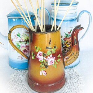May include: A vintage brown teapot with floral designs sits on a white doily with two blue and white teapots in the background. The teapot has a handle and a spout. The teapots are surrounded by white paper flowers on wooden sticks.