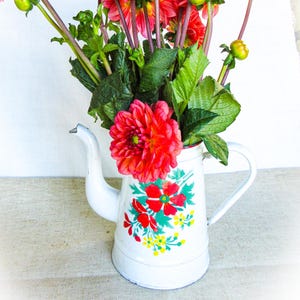 Op de afbeelding: Een witte emaille koffiepot met een bloemmotief van rode en gele bloemen en groene bladeren, gevuld met een boeket van levendige rode dahlia's op lange groene stelen met verschillende ongeopende groene bloemknoppen.