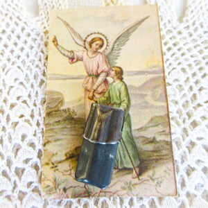 May include: A rectangular, dark gray metallic object with a smooth, polished finish is positioned over a vintage-style rectangular card. The card features a color illustration of an angel in a pink robe guiding a child in a green robe.