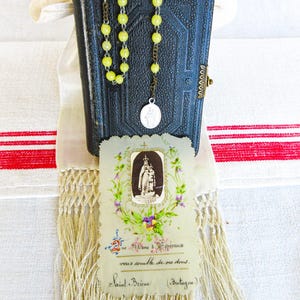 May include: A vintage rosary with yellow beads hangs over a small, dark blue book. A religious card with a floral border and text is in front of the book. The background includes a white cloth with red stripes and a fringed scarf.