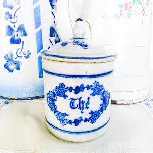 May include: A small, white ceramic tea jar with a lid, featuring blue floral designs and the word "The" in a decorative font. The jar is surrounded by other white and blue ceramic pieces.