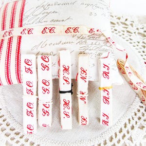 Könnte beinhalten: Vintage Bänder und ein Nadelkissen. Die weißen Bänder sind mit roten Buchstaben bedruckt, mit Initialen wie "E.C.", "F.O.", "H.S.", "N.S." und "R.J.". Das Nadelkissen hat rot-weiße Streifen.