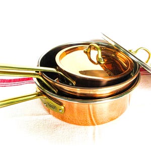 May include: A set of nested copper pans with polished brass handles and a matching copper lid. The pans feature a metallic finish and a small oval plaque on the side of the larger pan. The cookware is stacked together with the lid placed on top.