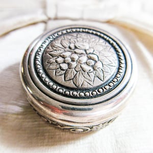 Può includere: Una scatola portaoggetti in argento con un design floreale. La scatola è rotonda e ha un bordo rialzato. Il design floreale è intricato e dettagliato.