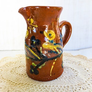 May include: A brown ceramic pitcher with a handle, painted with yellow, green, and black floral designs.