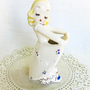 May include: A vintage ceramic figurine of a girl in a dress, with blonde hair and closed eyes. The off-white dress is decorated with purple floral accents and a scalloped hem. The figurine is standing on a lace doily.