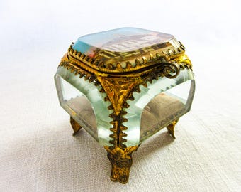 May include: A small, antique glass jewelry box with a hinged lid. The box has a rectangular shape with beveled glass sides and ornate gold-colored metal trim. The lid features a painted scene, and the box rests on small, decorative feet.