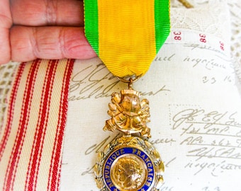 May include: A gold-toned medal featuring a central relief of a profile head surrounded by a blue enamel ring with the text REPUBLIQUE FRANCAISE. The medal is suspended from a yellow and green ribbon with a decorative gold-toned metal top piece.