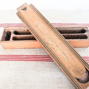 May include: A rectangular wooden pencil box with a sliding lid. The box features a light brown wood grain, a dark interior with two long compartments, and a decorative border pattern along the edges of the lid. The lid is partially slid open.