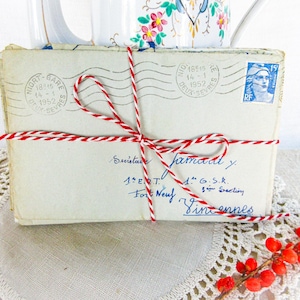 May include: A stack of vintage envelopes tied with red and white twine. The envelopes have stamps and handwritten addresses in blue ink. A sprig of red berries and a floral-patterned pitcher are in the background.