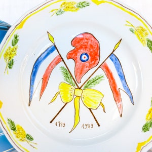 May include: A white ceramic plate with a yellow rim and a painted design of a red rooster with a blue eye, two crossed flags in red, white, and blue, and a yellow bow. The plate also has the dates "1789" and "1989" painted on it.