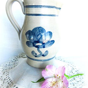 May include: A ceramic pitcher with a blue floral design and blue stripes around the top. The pitcher has a curved handle and a spout for pouring. A pink and purple flower rests on a white lace doily.