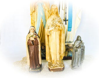 May include: A collection of religious statues. The statues depict figures in long robes, with one holding flowers. The statues are in various colors, including brown, beige, and silver. The background is a soft white.