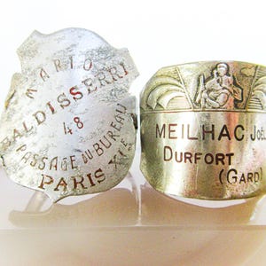 May include: Two vintage metal rings. One ring is shield-shaped with the text "MARIO BALDISSERRI 48 PASSAGE DU BUREAU PARIS XIE". The other ring is cylindrical with the text "MEILHAC Joel DURFORT (Gard)" and a relief design.