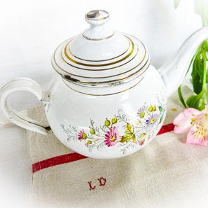 Könnte beinhalten: Weiße Emaille-Teekanne mit einem Blumendesign in Rosa, Gelb und Grün. Der Deckel und der Rand haben goldfarbene Akzente. Die Teekanne steht auf einem Leinentuch mit rotem Streifen und einer rosa Blume.