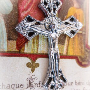 Puede incluir: Un colgante de crucifijo plateado detallado con una figura de Jesucristo. La cruz tiene un dise&ntilde;o de filigrana ornamentado y se encuentra sobre un fondo de papel envejecido con texto en franc&eacute;s y una ilustraci&oacute;n religiosa. Un art&iacute;culo religioso.