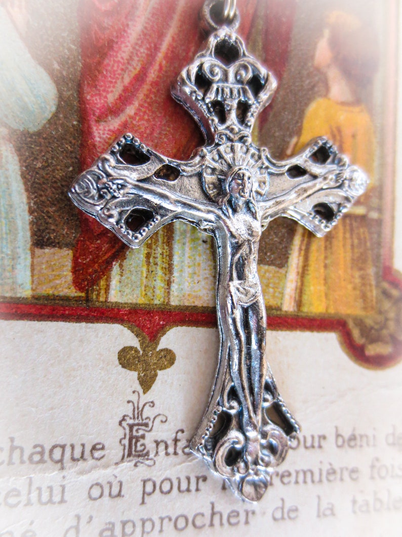 Puede incluir: Un colgante de crucifijo plateado detallado con una figura de Jesucristo. La cruz tiene un dise&ntilde;o de filigrana ornamentado y se encuentra sobre un fondo de papel envejecido con texto en franc&eacute;s y una ilustraci&oacute;n religiosa. Un art&iacute;culo religioso.