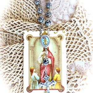 Puede incluir: Un rosario con cuentas plateadas y un colgante de cruz detallado. El rosario cuelga sobre una tarjeta religiosa que representa a Jes&uacute;s con dos ni&ntilde;os. El fondo incluye un tapete de encaje de ganchillo.