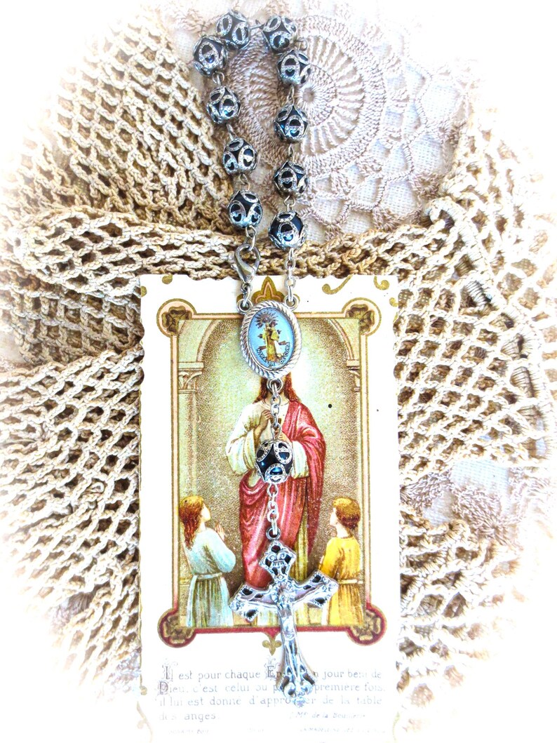 Puede incluir: Un rosario con cuentas plateadas y un colgante de cruz detallado. El rosario cuelga sobre una tarjeta religiosa que representa a Jes&uacute;s con dos ni&ntilde;os. El fondo incluye un tapete de encaje de ganchillo.