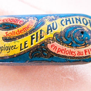Op de afbeelding: Een vintage blauwe en gele tinnen doos met sierlijke gouden letters die "Pour coudre solidement employez LE FIL AU CHINOIS en pelotes au FIL D'OR" luidt.