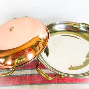May include: Two copper and stainless steel pans with brass handles. One pan is copper with a shiny finish, and the other is stainless steel. Both pans are round and sit on a red and white striped surface.