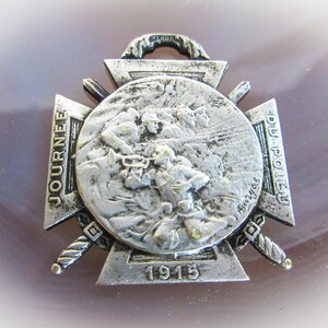 May include: A silver-toned medal with a cross design. The center of the medal features a relief image of soldiers in battle. The medal is inscribed with the text "JOURNEE DU POILU" and "1915".
