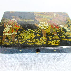 May include: Black rectangular box with a hinged lid, featuring a painted scene of figures in traditional attire. The artwork includes gold and green accents, with the word "DEPOSE" visible on the side.