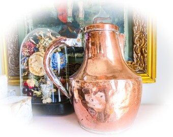 May include: A copper pitcher with a rounded body and a handle. The pitcher has a shiny, reflective surface and appears to be antique.